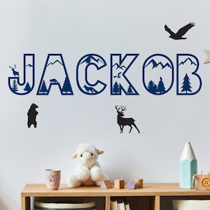 Könnte beinhalten: Ein blauer und weißer Wandaufkleber mit dem Namen "JAKOB" in großen Buchstaben. Die Buchstaben sind mit einer Berg- und Waldlandschaft verziert. Es gibt auch Bilder von einem Hirsch, einem Bären und einem Vogel.
