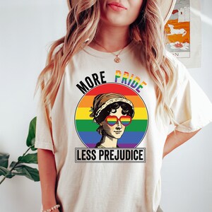 May include: A white t-shirt with a rainbow circle graphic featuring a woman wearing heart-shaped sunglasses. The text "More Pride Less Prejudice" is printed below the circle.
