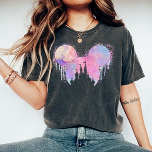 May include: A dark gray t-shirt with a watercolor design of Mickey Mouse ears and the silhouette of Cinderella's castle. The design is in shades of pink, purple, and blue.