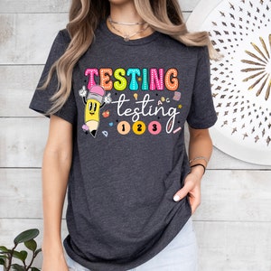 May include: A dark gray t-shirt with the text "TESTING testing 1 2 3" in colorful letters. A cartoon pencil with a smiling face is next to the text.