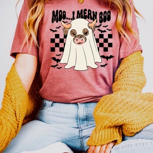 May include: A pink t-shirt with a graphic of a Highland cow dressed as a ghost. The text "Moo...I mean Boo" is printed on the shirt.