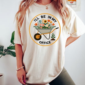 May include: A white t-shirt featuring a graphic of a wheelbarrow full of flowers and the text "I'll be in my office".