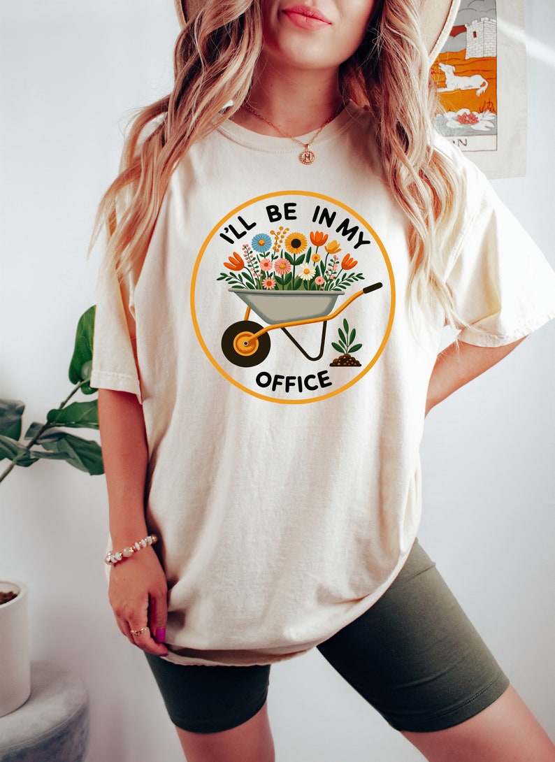 May include: A white t-shirt featuring a graphic of a wheelbarrow full of flowers and the text "I'll be in my office".