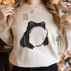 May include: A cream-colored sweatshirt with a black frog illustration and a red stamp on the front.
