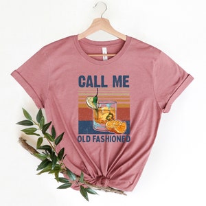 May include: A pink t-shirt with the text "CALL ME OLD FASHIONED" in white and a vintage-style illustration of an old fashioned cocktail with a lime wedge, orange slices, and a green chili pepper.
