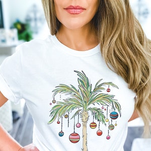 May include: White t-shirt with a graphic of a palm tree decorated with colorful ornaments. The palm tree is drawn in a cartoon style with green leaves and a brown trunk.