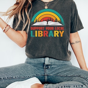 May include: A dark gray t-shirt with a vintage-style graphic of a book with a rainbow arch and trees. The text on the shirt reads "Support Your Local Library".