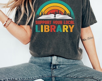 May include: A dark gray t-shirt with a vintage-style graphic of a book with a rainbow arch and trees. The text on the shirt reads "Support Your Local Library".