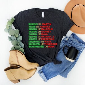 May include: A dark grey t-shirt with a red and green graphic print that reads "Dream like Martin, Lead like Harriet, Fight like Malcolm, Think like Garvey, Write like Maya, Build like Madam C.J., Speak like Frederick, Educate like W.E.B., Believe like Thurgood, Challenge like Rosa".