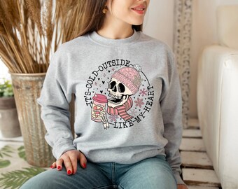 May include: Gray sweatshirt with a graphic of a skull wearing a pink hat and scarf, holding a cup of coffee. The text on the sweatshirt reads "It's cold outside like my heart."