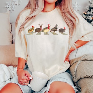 May include: A white t-shirt with a graphic of five quail wearing Santa hats and Christmas lights walking across a crosswalk.