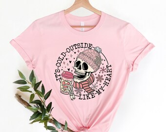 May include: A light pink t-shirt featuring a graphic of a skull wearing a pink knit hat and scarf, holding a coffee cup. The text "IT'S COLD OUTSIDE LIKE MY HEART" is printed around the skull in a circular design. The shirt is hanging on a wooden hanger.