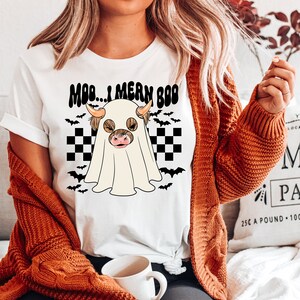 May include: White t-shirt with a black and white checkered background and a ghost with a brown and white cow head inside. The text "MOD... I MEAN BOO" is above the ghost.