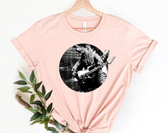 May include: A pink t-shirt with a black and white graphic of a Godzilla-like creature playing an electric guitar. The graphic is in a circle with a distressed background.