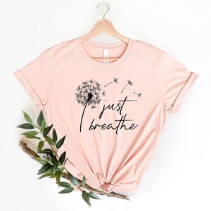Op de afbeelding: Een lichtroze T-shirt met een zwarte paardenbloem afbeelding en de tekst "just breathe".