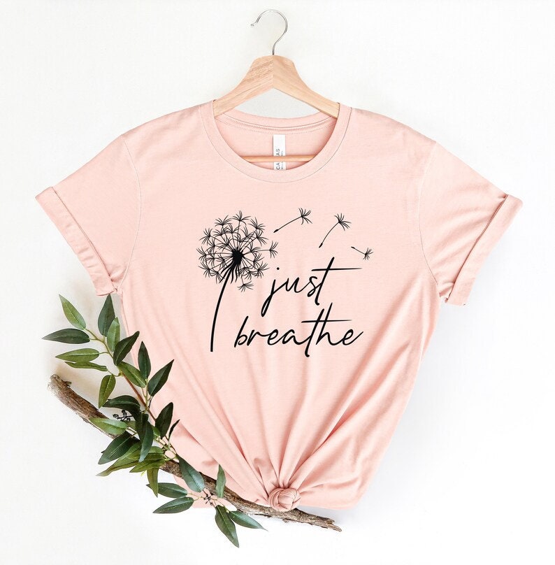 Op de afbeelding: Een lichtroze T-shirt met een zwarte paardenbloem afbeelding en de tekst "just breathe".