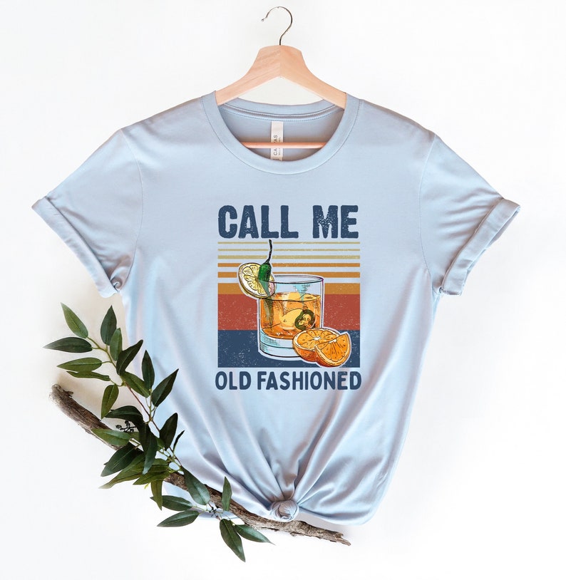 May include: Light blue t-shirt with a vintage-style graphic of a cocktail glass with a lemon wedge, orange slices, and a chili pepper. The text "Call Me Old Fashioned" is printed below the image.