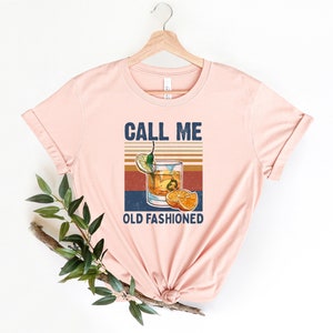 May include: A pink t-shirt with the text "Call Me Old Fashioned" and a vintage-style illustration of an old fashioned cocktail with orange slices and a lime wedge.