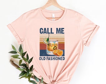 May include: A pink t-shirt with the text "Call Me Old Fashioned" and a vintage-style illustration of an old fashioned cocktail with orange slices and a lime wedge.