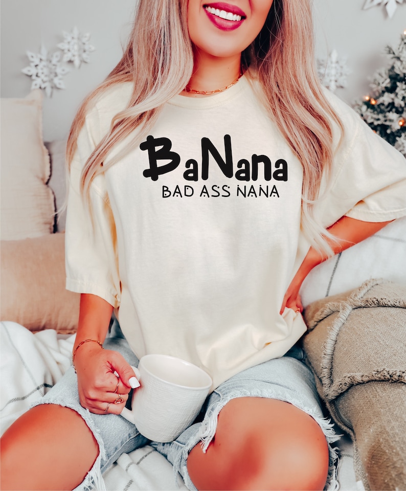 May include: A woman wearing a light yellow t-shirt with the text "BaNana BAD ASS NANA" in black. She is sitting on a couch and holding a white mug.