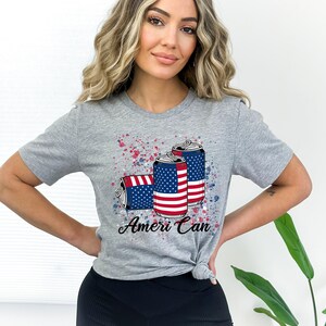 May include: A gray t-shirt with a graphic of three red, white, and blue American flag-themed cans. The text "Ameri Can" is printed below the cans.
