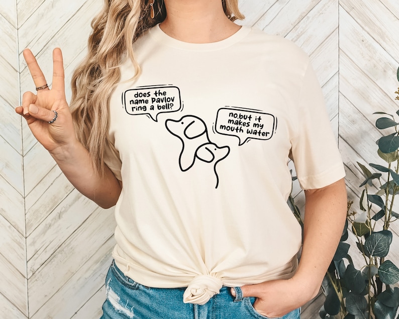May include: A white t-shirt with a black illustration of two dogs. The text on the shirt says "does the name pavlov ring a bell?" and "no but it makes my mouth water."