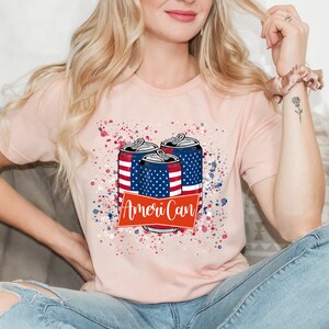 May include: A pink t-shirt with a graphic of three aluminum cans with the American flag design. The cans are surrounded by red, white, and blue paint splatters. The text "Ameri-Can" is printed in red on a white banner.