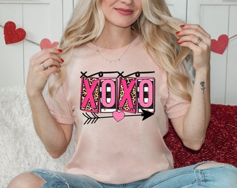 May include: Pink t-shirt with a graphic of the word "XOXO" in pink letters with a leopard print and black and white stripes. The letters are surrounded by a black outline and a string of lights. There is a pink heart and arrows on either side of the letters.