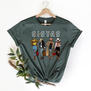May include: A green t-shirt with the text "SISTAS" in white with colorful dots. The shirt features an illustration of four women wearing hats and stylish outfits.