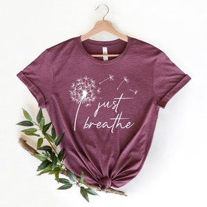 Op de afbeelding: Een maroon t-shirt met een witte paardenbloem afbeelding en de tekst "just breathe" gedrukt op de voorkant.