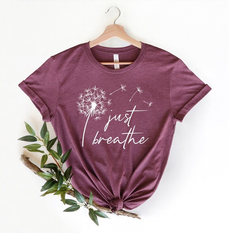 Op de afbeelding: Een maroon t-shirt met een witte paardenbloem afbeelding en de tekst "just breathe" gedrukt op de voorkant.