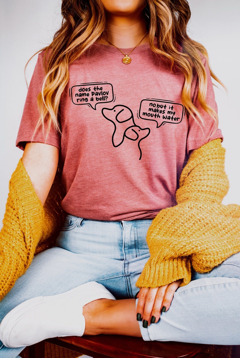 May include: A pink t-shirt with a black illustration of two dogs and speech bubbles. The text in the speech bubbles reads "does the name pavlov ring a bell?" and "no, but it makes my mouth water".