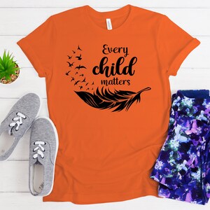 May include: Orange t-shirt with the text "Every child matters" in black, along with a black feather and birds graphic. The shirt is paired with gray sneakers and patterned leggings. A small potted plant is also visible.