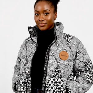 May include: A black and white patterned puffer jacket with a high collar. The jacket features a geometric design and a brown leather patch with the text "BIBILUXE". The model is wearing a black turtleneck.