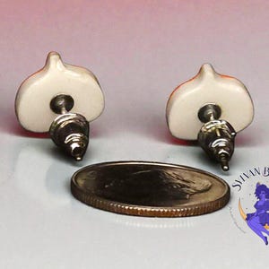 Peut inclure: Une paire de boucles d'oreilles &agrave; clous avec un motif blanc et orange, avec des tiges argent&eacute;es. Une pi&egrave;ce de monnaie est &agrave; c&ocirc;t&eacute; des boucles d'oreilles. Les boucles d'oreilles sont sur une surface claire. Les boucles d'oreilles sont de Sylvan Baubles.