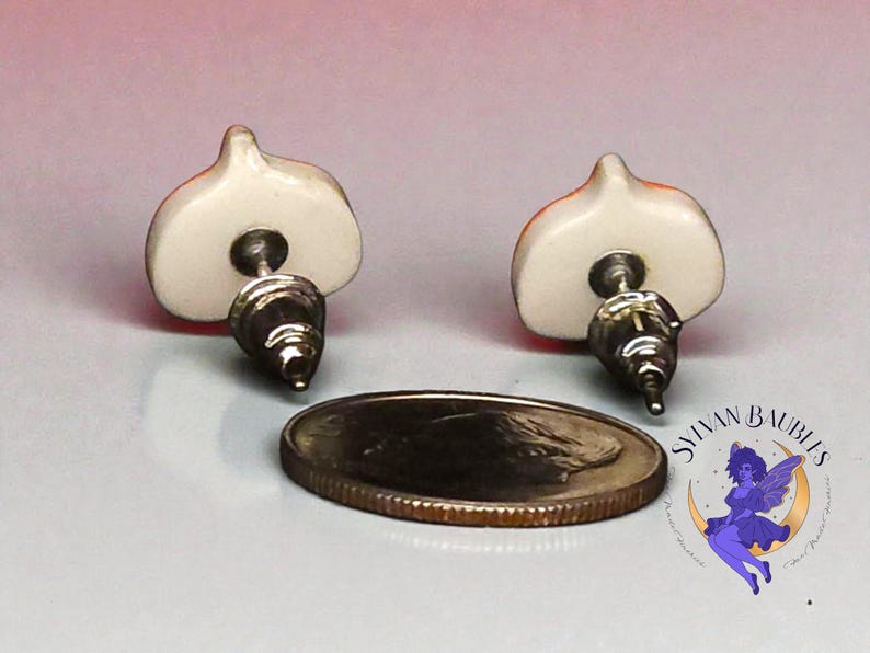 Peut inclure: Une paire de boucles d'oreilles &agrave; clous avec un motif blanc et orange, avec des tiges argent&eacute;es. Une pi&egrave;ce de monnaie est &agrave; c&ocirc;t&eacute; des boucles d'oreilles. Les boucles d'oreilles sont sur une surface claire. Les boucles d'oreilles sont de Sylvan Baubles.