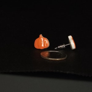 Peut inclure: Boucles d'oreilles &agrave; clous en forme de citrouille orange et une boucle d'oreille unique avec un motif blanc et orange. Les boucles d'oreilles sont pr&eacute;sent&eacute;es sur une surface noire &agrave; c&ocirc;t&eacute; d'une pi&egrave;ce de monnaie.