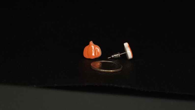 Peut inclure: Boucles d'oreilles &agrave; clous en forme de citrouille orange et une boucle d'oreille unique avec un motif blanc et orange. Les boucles d'oreilles sont pr&eacute;sent&eacute;es sur une surface noire &agrave; c&ocirc;t&eacute; d'une pi&egrave;ce de monnaie.
