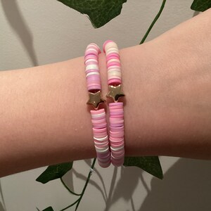 Może przedstawiać: Dwa bransoletki z koralików w kolorze różowym i białym z zawieszkami w kształcie złotej gwiazdki. Bransoletki są ułożone na nadgarstku osoby.