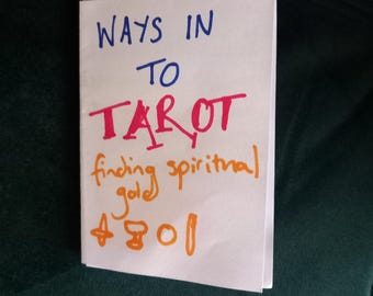Pode incluir: Um cartão branco com as palavras "Ways in to Tarot finding spiritual gold" escritas em marcador azul, vermelho e laranja. O cartão também tem os símbolos dos quatro naipes do tarot: varinhas, copas, pentáculos e espadas.