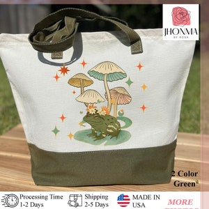K&ouml;nnte beinhalten: Eine wei&szlig;e Canvas-Tragetasche mit gr&uuml;nem Boden. Die Tasche zeigt eine fantasievolle Illustration eines Frosches mit einer Blumenkrone, der auf einem Seerosenblatt sitzt, umgeben von Pilzen und Sternen. Der Text "JHONMA BY ROSA" ist in der oberen rechten Ecke der Tasche aufgedruckt.