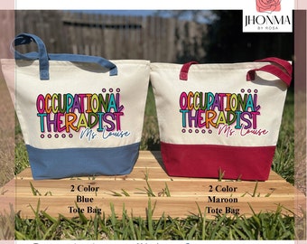 May include: Two canvas tote bags with a two-toned design. The top half of each bag is white with the text "Occupational Therapist" in colorful letters. The bottom half of the bag is a solid color, one is blue and the other is maroon. The text "Ms. Louise" is printed below the main text.
