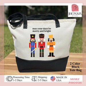 May include: A white canvas tote bag with black handles and a black bottom. The bag has a design of three nutcracker figures with the text "may your days be merry and bright". The bag is labeled "2 Color Black Tote Bag".