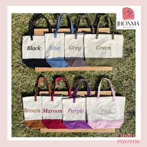 May include: Canvas tote bags in various colors: black, blue, grey, green, brown, maroon, purple, and pink. Each bag has a contrasting bottom panel and matching handles. The names of the colors are printed on the bags.
