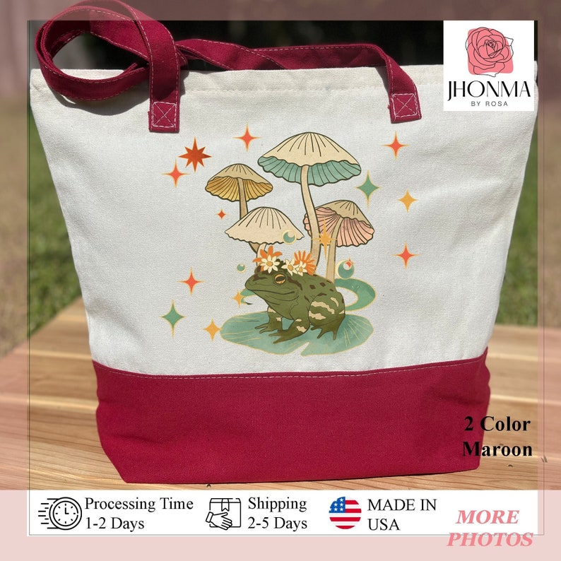 K&ouml;nnte beinhalten: Eine wei&szlig;e Canvas-Tragetasche mit einem maroonfarbenen Boden. Die Tasche zeigt ein fantasievolles Design mit einem Frosch, der eine Blumenkrone tr&auml;gt und unter einer Gruppe von Pilzen sitzt. Das Design ist von Sternen und Glitzer umgeben.
