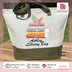 Op de afbeelding: Een witte canvas draagtas met groene handvatten en een groene bodem. De tas heeft een ontwerp met een stapel boeken waar een konijn uit piept. De tekst "Ashley Library Bag" is op de tas gedrukt.