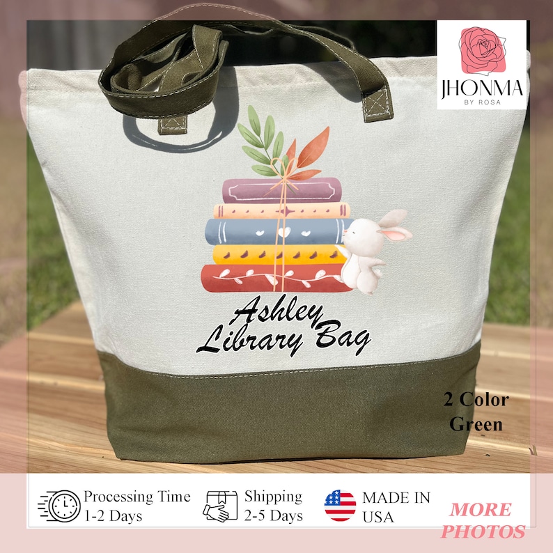 Op de afbeelding: Een witte canvas draagtas met groene handvatten en een groene bodem. De tas heeft een ontwerp met een stapel boeken waar een konijn uit piept. De tekst "Ashley Library Bag" is op de tas gedrukt.