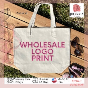 Pu&ograve; includere: Una borsa tote in tela naturale con il testo "WHOLESALE LOGO PRINT" in lettere rosa. La borsa &egrave; su una superficie di legno con un nastro rosa, occhiali da sole e trucco.