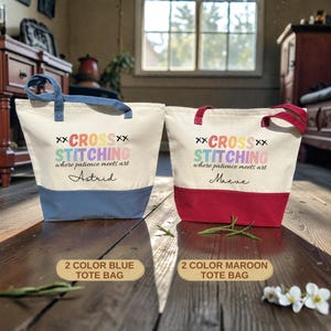 May include: Two canvas tote bags, one with a blue base and the other with a maroon base. Both bags have the text "CROSS STITCHING where patience meets art" printed on them. The blue bag is labeled "Astrid" and the maroon bag is labeled "Maeve".