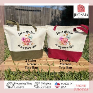 May include: Two tote bags with a white canvas top and a colored bottom. The top of the bag has a design of a ball of yarn with needles and flowers. The text on the bag reads "I'm a Hooker in my spare time". The bag on the left has a green bottom and the bag on the right has a maroon bottom.